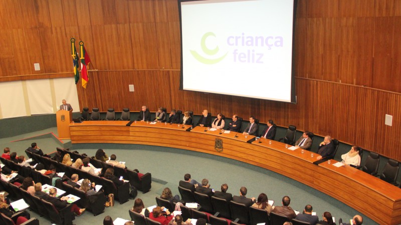 Foto tirada da parte de cima do auditório mostra os participantes do evento atrás de uma bancada, prestando a atenção na fala do secretário Gabbardo, que está de pé atrás de um púlpito, com um microfone à sua frente. 