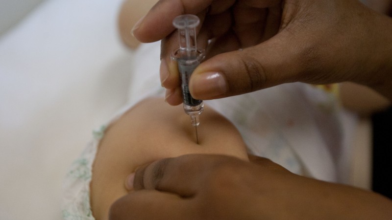 A imagem mostra duas mãos vacinando a perna de um bebê. 