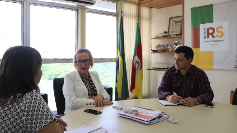Arita conversa com o gerente de planejamento Ronaldo Carbonari e a gerente-adjunta de comunicação, Maria Elizabete Aguiar. Os três estão sentados atrás de uma mesa oval. 