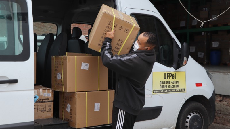 Um homem carregando uma caixa para dentro de uma van. A van tem o adesivo da Universidade Federal de Pelotas.