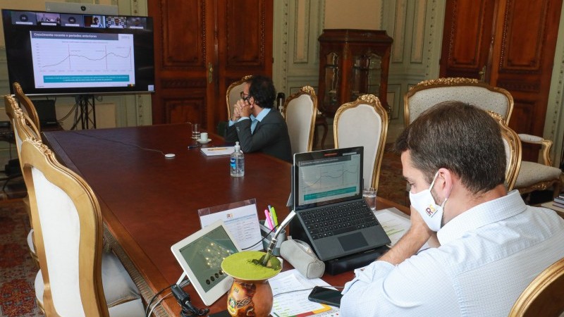 Eduardo Leite olha para o gráfico que está no computador a sua frente enquanto o outro participante da reunião olha para o mesmo gráfico que está num telão a sua frente. 