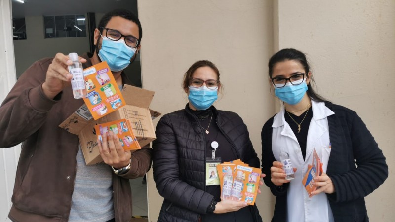 Três profissionais (um homem e duas mulheres, todos de máscara) com os materiais  (folhetos e álcool gel) para fazer a entrega. 