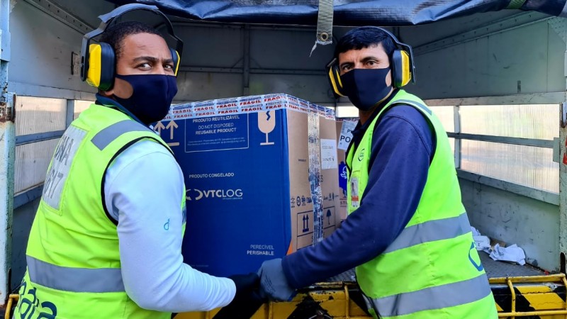 Dois funcionários (de máscara) olham para a frente e carregam uma caixa grande. 