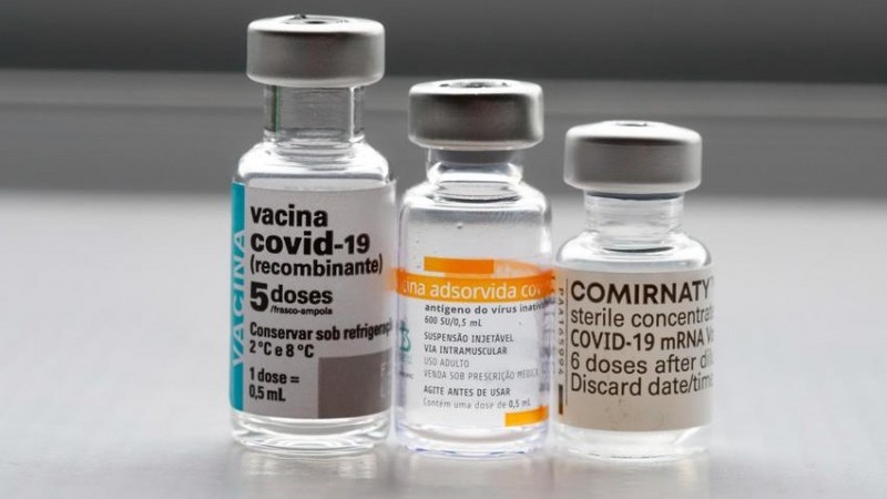 A imagem mostra três frascos de vacinas contra a covid em cima de uma superfície. 