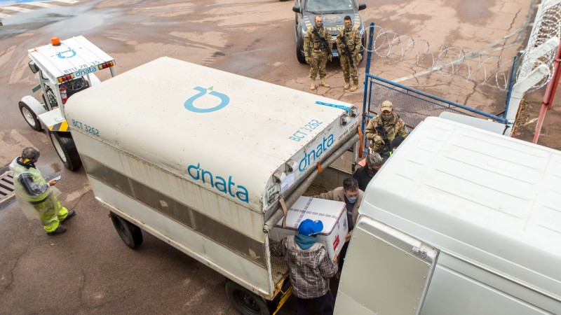 Dois funcionários tiram uma caixa, do reboque, grande isopor com vacinas para botar na caminhonete. Três militares acompanham a cena.  