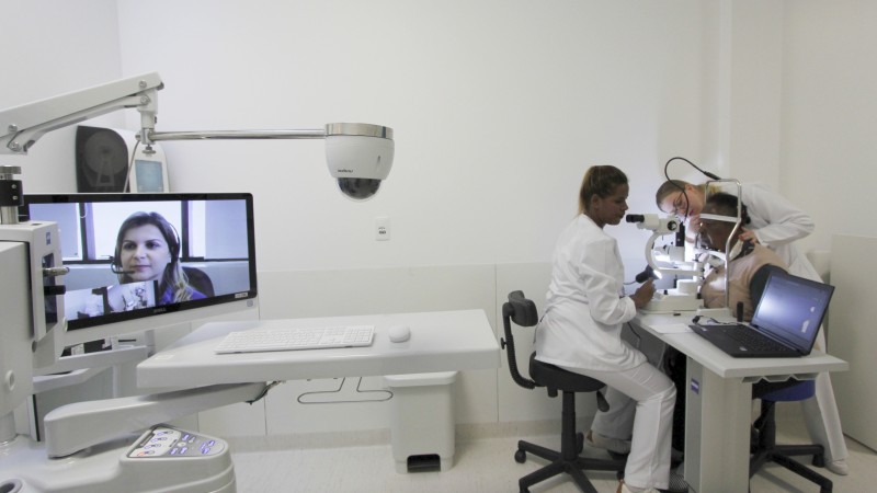 Duas médicas atendem uma senhora. Uma doutora acompanha o atendimento através de uma tela de computador. 