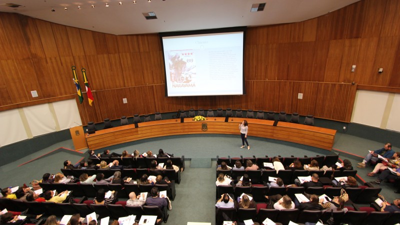 A imagem mostra o público sentado, assistindo atento a fala da palestrante que, com o microfone na mão, olha para o telão. 