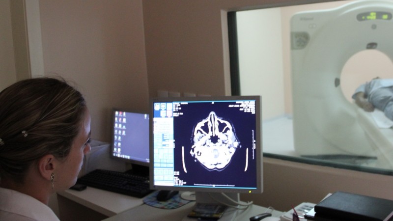 Médica olha para a tela do computador. Na tela, a imagem é o resultado de um exame. No fundo, um paciente está deitado na maca. 