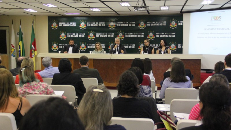 Secretário Gabbardo, sentado atrás de uma mesa, discursa com um microfone nas mãos. Ele está no meio de quatro pessoas (um homem e três mulheres).