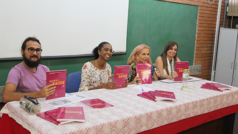 Autores (um homem e três mulheres), sentados atrás de uma mesa, seguram o livro. 