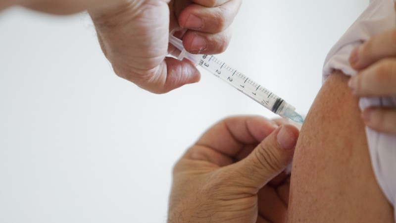 Imagem mostra as mãos aplicando uma vacina no braço de outra pessoa. 