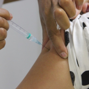duas mãos de umaa pessoa aplicando a vacina com aparelho descartável em um ombro de uma pessoa com blusa branca e bola pretas.